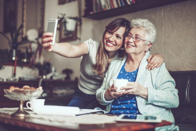 Grandmother and granddaughter take a selfie on a mobile phone together. They are smiling at the camera.