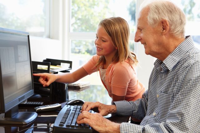 A young girl and older man use a computer. The young girl is pointing at the monitor. 