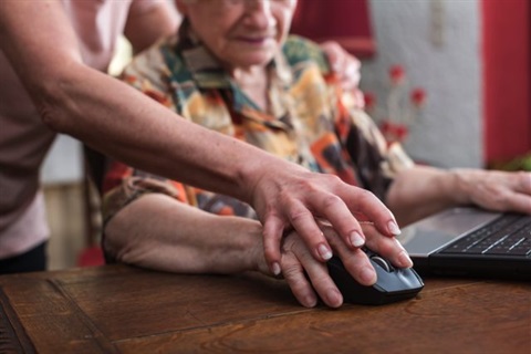 Close up of someone helping an older person to use a computer. Their hands rest on a computer mouse.