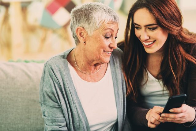 An older woman and young woman sit on a couch. The young woman is showing the older woman how to use a mobile phone.